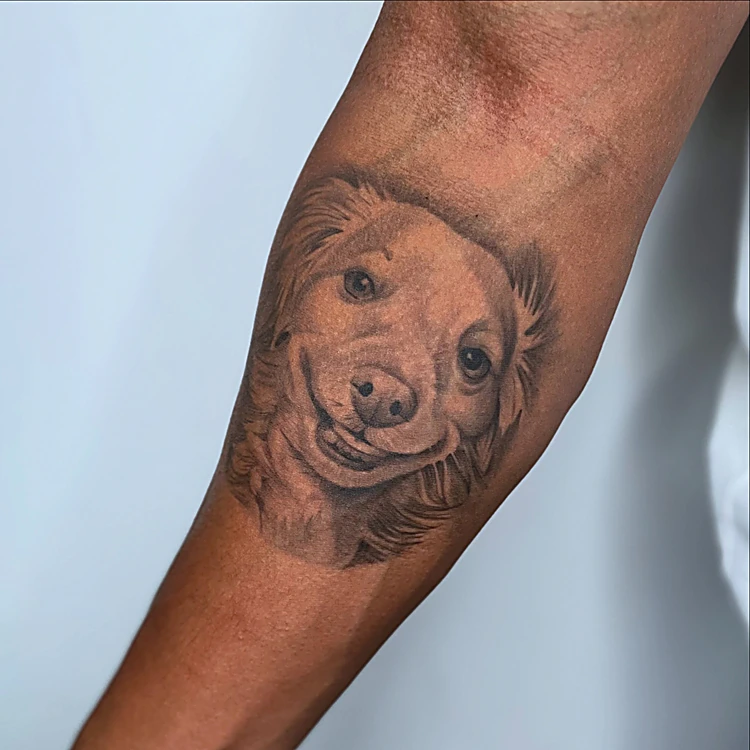 Retrato realista de un perro de pelo claro sonriendo, en negro y gris sobre el antebrazo